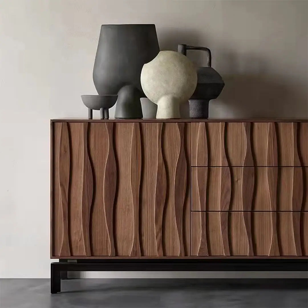 close-up view of a modern sideboard made from walnut wood, featuring a unique wave-like texture on its doors and drawer fronts. The sideboard has a sleek and elegant design with three drawers on the right side and two doors on the left. The surface of the sideboard is adorned with decorative vases and sculptures in various shapes and sizes, adding an artistic touch to the overall look. The piece stands on sturdy black metal legs, providing a contrast to the warm tones of the walnut wood.