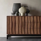 close-up view of a modern sideboard made from walnut wood, featuring a unique wave-like texture on its doors and drawer fronts. The sideboard has a sleek and elegant design with three drawers on the right side and two doors on the left. The surface of the sideboard is adorned with decorative vases and sculptures in various shapes and sizes, adding an artistic touch to the overall look. The piece stands on sturdy black metal legs, providing a contrast to the warm tones of the walnut wood.