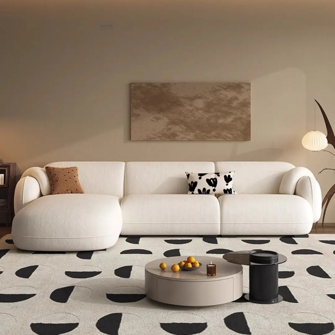 Stylish living room featuring a large, cream-colored L-shaped sofa with rounded edges. The sofa is complemented by matching pillows, one with a brown and the other with a black and white pattern. The room includes a cozy armchair of the same color and a round, beige ottoman.