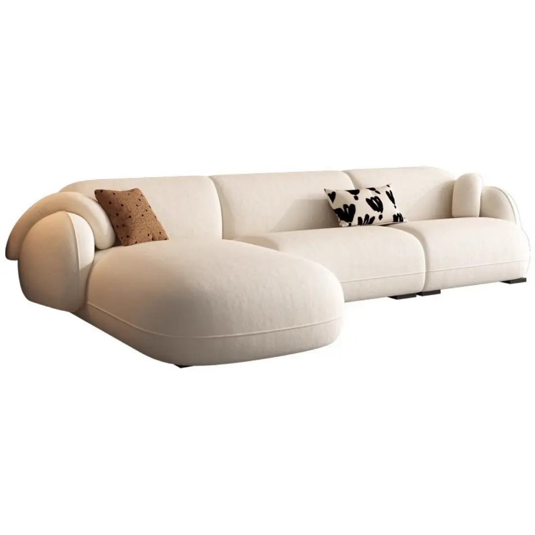 L-shaped corner tofu sofa in a neutral beige color. The sofa has a clean, minimalist design with a left-side chaise lounge. The seat cushions are thick and plush, providing a comfortable seating experience. The sofa is accented with two throw pillows, one in a light brown with a subtle pattern and the other in black and white with abstract designs. The overall style is contemporary and inviting, perfect for a modern living room setting.