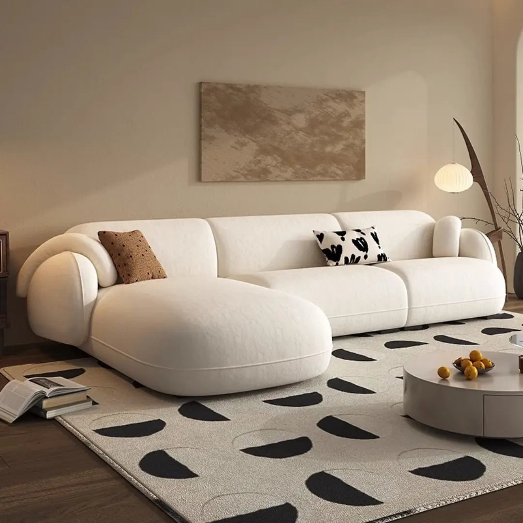 The sofa is upholstered in a cream-colored fabric and includes plush, rounded armrests. It has a chaise lounge section on the left, topped with a brown throw pillow, and the main seating area includes a black and white patterned cushion. The room is decorated with a minimalist aesthetic, including a geometric rug in black and cream, a round coffee table with a few decorative items, and a modern floor lamp.