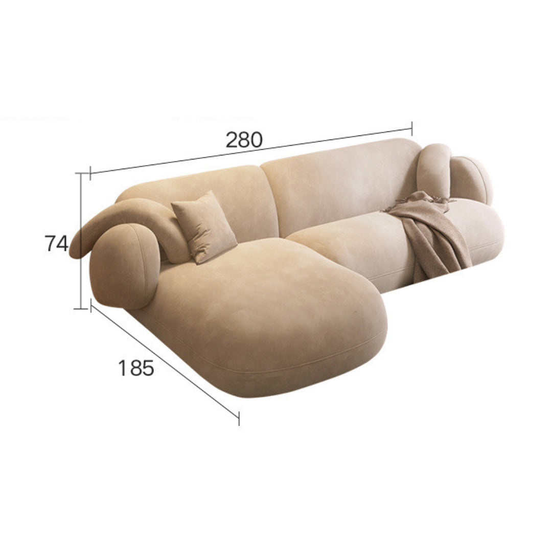 Tofu_Sofa_280_cm is upholstered in a cream-colored fabric and includes plush, rounded armrests. It has a chaise lounge section on the left, topped with a brown throw pillow, and the main seating area includes a black and white patterned cushion.