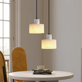 Two alabaster white marble pendant lights hanging above a modern round dining table with mustard yellow chairs, styled in a sunlit interior with large windows.