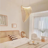 A well-lit, cozy bedroom featuring a unique, sculptural LED chandelier with a sweeping, curved design, suspended from the ceiling. The room is styled with neutral colors, including beige and soft white, for a tranquil atmosphere. The bed has a rattan headboard, and is dressed in white and beige linens, with a polka-dotted throw pillow adding a touch of pattern. To the side, a modern white chair and a small wooden side table with a potted plant contribute to the room's organic feel.