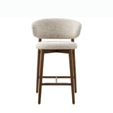 The stool has a modern design, featuring a cushioned seat and a curved backrest, both upholstered in a light, textured fabric. The frame and legs are made of wood, providing a sturdy and natural look. A horizontal wooden beam supports the backrest, enhancing the stool's aesthetic and structural integrity.