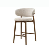 The stool features a cushioned seat and a rounded backrest, both upholstered in a light, textured fabric. The legs and frame are made of wood, providing a sturdy and natural look. The overall design is minimalist and contemporary, making it an ideal addition to any kitchen counter, 