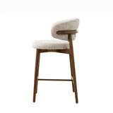 The image shows a single, stylish high stool viewed from the side. The stool features a contemporary design with a cushioned seat and a curved backrest, both upholstered in a light, textured fabric. The legs and frame of the stool are made of wood, providing a natural and sturdy base. The design is minimalistic, with clean lines and a modern aesthetic, making it suitable for various interior settings such as a kitchen counter or bar area.