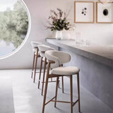 modern kitchen or bar area featuring a sleek counter and four high stools. The stools have a contemporary design with cushioned seats and curved backrests upholstered in a light, textured fabric. The legs of the stools are made of wood, adding a natural touch to the clean, minimalist aesthetic. The counter is light grey, and on top of it, there are a few decorative items, including a vase with flowers and some glassware.