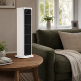 A white tower fan with a black front grille stands on a round wooden side table in a cozy living room setting next to a green sofa.