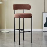 A Brown Whittington Teddy Delightful Bar Stool featuring a white textured two-tier seat with a vertical backrest, supported by a minimalist black metal frame, positioned in a modern interior with light flooring and a white floor lamp in the background.