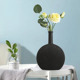 A matte black ribbed vase with fresh yellow roses and greenery, accompanied by a small clear bottle with a single green stem, presented on a white tray against a soft blue wall.