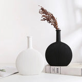 Two minimalist ceramic vases on a table, one matte white with vertical ribbing, the other matte black with a bouquet of dried flowers, against a clean, neutral background.