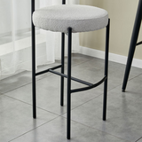 Close-up view of a Whittington Teddy Delightful Bar Stool featuring a grey plush seat and a sleek black metal frame, positioned on a grey tile floor against a minimalist white curtain backdrop.