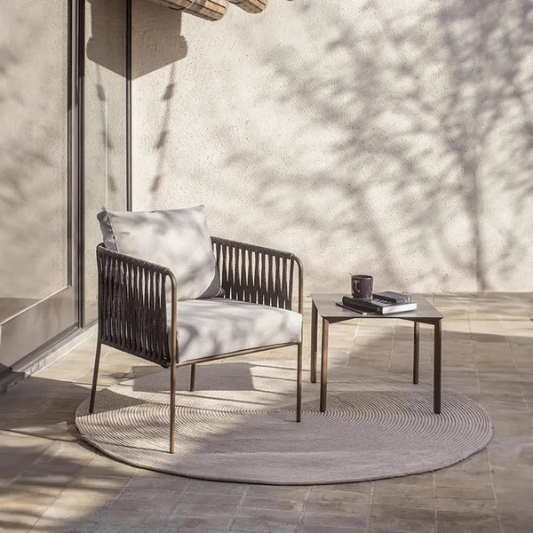 Minimalist outdoor patio setup featuring a modern lounge chair with gray cushions, paired with a sleek metal side table. The furniture is set on a neutral-toned circular rug, with soft natural light and tree shadows creating a serene atmosphere against the textured wall.