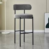 A Luxury  Whittington Teddy Delightful Bar Stool featuring a white textured two-tier seat with a vertical backrest, supported by a minimalist black metal frame, positioned in a modern interior with light flooring and a white floor lamp in the background.