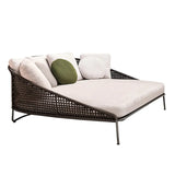 A modern outdoor daybed with a woven rattan frame, featuring a wide, cushioned seat and a variety of throw pillows in white, beige, and green. The daybed is elevated on slim metal legs, providing a sleek and comfortable lounging option for outdoor spaces.