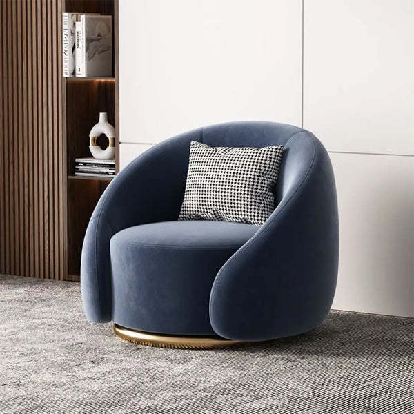 A deep blue velvet swivel armchair with a rounded, enveloping backrest and a gold-tone base. The chair is paired with a black-and-white houndstooth pillow and set against a contemporary white wall with wooden accents.