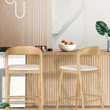 Raw wood bar stools in a modern kitchen island setting, featuring a mid-century bar stool design with ash wood frame, upholstered white seat and light wood wall panelling.