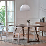  The table has a rectangular wooden top with angled legs forming an open triangular shape, providing a contemporary look. Surrounding the table are white chairs with curved backrests and woven seats, adding a touch of contrast and lightness to the setting. The table is placed on a patterned rug, and a white pendant lamp hangs overhead, enhancing the modern aesthetic. A small potted plant and neatly stacked plates adorn the table. 