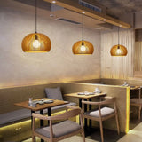 A cozy restaurant interior illuminated by three golden-hued wooden pendant lights, which are shaped like pumpkins with slatted designs that cast a warm glow over the dining area. The lights hang above wooden tables paired with comfortable chairs and a bench, set against a backdrop of minimalist wooden walls and a softly textured beige wall. The ambiance is welcoming and stylish, perfect for a relaxed dining experience.