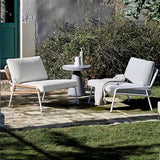 An outdoor lounge set with two armchairs and a small table, featuring sleek white cushions and a combination of metal and rattan frames, set against a natural stone patio with a backdrop of greenery and a gray wall.