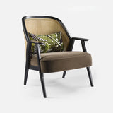 Side angle view of the Qienn Medieval Rattan Armchair featuring a natural rattan backrest, dark wooden frame, and olive-green velvet seat cushion, complemented by a decorative pillow with a tropical leaf pattern.