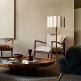 A cozy living room setup featuring a wooden table lamp with a cylindrical, pleated shade, standing next to a mid-century wooden armchair. A wooden round coffee table with books and rustic decor items completes the warm, inviting scene.