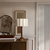 Elegant interior setup showing a classic wooden floor lamp with a tall, sleek base and a cream-colored pleated shade, positioned on a dark wooden sideboard. The sideboard also hosts a stack of books and a framed art piece, enhancing a refined home decor ambiance, beside an arched mirror reflecting a branch arrangement.