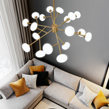 The image displays a modern Tadley 15-lights chandelier, featuring a golden structure with spherical white shades arranged in a dynamic, sprawling form. The chandelier hangs above a contemporary gray sofa adorned with gold and black cushions, creating a striking contrast against a dark gray wall, enhancing the room's modern elegance.