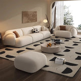  a modern, stylish living room featuring a large, cream-colored L-shaped sofa with rounded edges. The sofa is complemented by matching pillows, one with a brown and the other with a black and white pattern. The room includes a cozy armchair of the same color and a round, beige ottoman. The space is adorned with a light-colored rug featuring black abstract patterns, a round coffee table, and minimalistic decor including a floor lamp