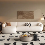 , stylish living room featuring a large, cream-colored L-shaped sofa with rounded edges. The sofa is complemented by matching pillows, one with a brown and the other with a black and white pattern. The room includes a cozy armchair of the same color and a round, beige ottoman.