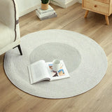 A spacious gray round rug with a concentric circular texture is laid out on a wooden floor, complementing the minimalistic decor of the room. An open book and a white cup on a small plate rest on the rug, creating a cozy and inviting reading nook next to an off-white armchair and a light wooden side table with plants and books.