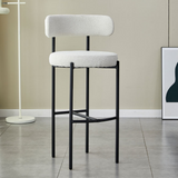 A sleek Whittington Teddy Delightful Bar Stool featuring a white textured two-tier seat with a vertical backrest, supported by a minimalist black metal frame, positioned in a modern interior with light flooring and a white floor lamp in the background.