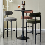 A stylish arrangement of Whittington Teddy Delightful Bar Stools in varied textures of grey, rose, and green, surrounding a sleek black round table with two bottles of wine, set in a modern, light-filled room.