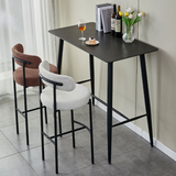 A contemporary dining setup featuring two Whittington Teddy Delightful Bar Stools in rose and grey, paired with a black rectangular high table adorned with a glass of wine, a dessert, books, and a vase of fresh tulips, in a chic, airy room.