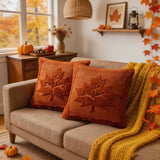 Two square orange-brown throw pillows with raised leaf designs on a beige sofa in a cozy autumn living room setting with a yellow knit blanket, pumpkins, and fall decorations.