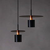 Two elegant pendant lights with a sleek design, featuring flat, circular shades in matte black, complemented by cylindrical bodies and interior accents in a copper finish. These lights are suspended against a dark gray background, highlighting their modern and sophisticated appearance. Each light hangs from a thin, black cable, adding to the overall minimalist and contemporary aesthetic.