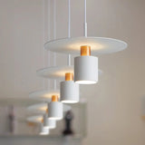 A group of modern pendant lights featuring a harmonious arrangement of flat, disc-shaped shades and cylindrical elements, all in a white and copper color scheme. The lights hang at various heights, creating a visually appealing installation suitable for stylish interiors. Each cylindrical element is centrally positioned beneath its respective disc, emitting soft light that enhances the ambiance of the space.