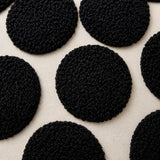 Close-up of a black textured circular fabric or rug pattern on a warm off-white woven background.