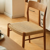 A modern, minimalist Qeinn Ratten Naas dining chair with a polished wooden frame and a natural rattan seat, set against a light wood floor and a simple, elegant home decor backdrop.