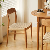 A warmly lit dining area features the Qeinn Ratten Naas dining chair with a sleek wooden frame and woven rattan seat, complementing a round wooden table with a cup of coffee and a white orchid in the background, near a window with sheer curtains.