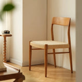 Elegant Qeinn Ratten Naas dining chair with a natural wood finish and woven rattan seat, positioned in a warmly lit room next to a small wooden side table.